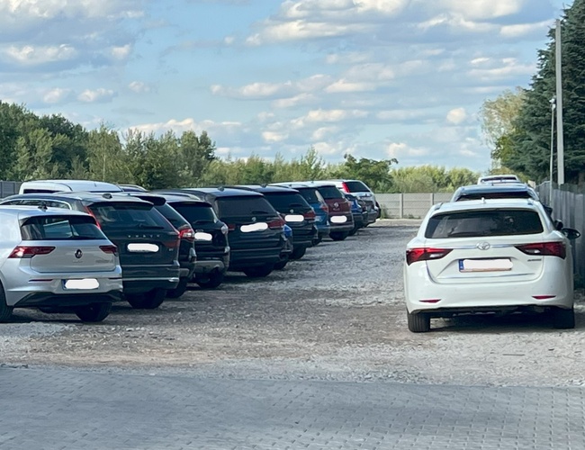 PRO-PARK – Parking z samochodami. Teren jest żwirowy i otwarty. Po lewej stronie widoczne są drzewa.