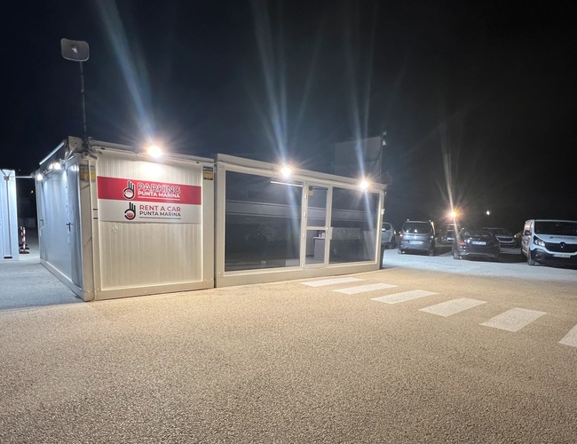 Parkering Punta Marina - Moderne kontorcontainer opplyst av lamper. Flere parkerte biler ved siden av en godt merket vei.