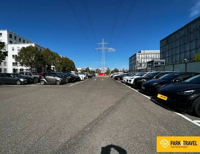 Parking Park2Travel Valet Outdoor - Szeroki plac z zaparkowanymi samochodami po obu stronach. Teren otoczony jest budynkami i ogrodzeniem.