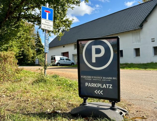 W centrum znak parking, w tle zaparkowany samochód budynek i drzewa.