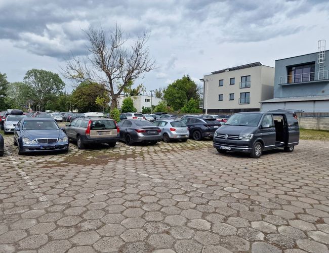 Widok samochodów zaparkowanych na parkingu z samochodem transferowym. Utwardzony kostką teren.
