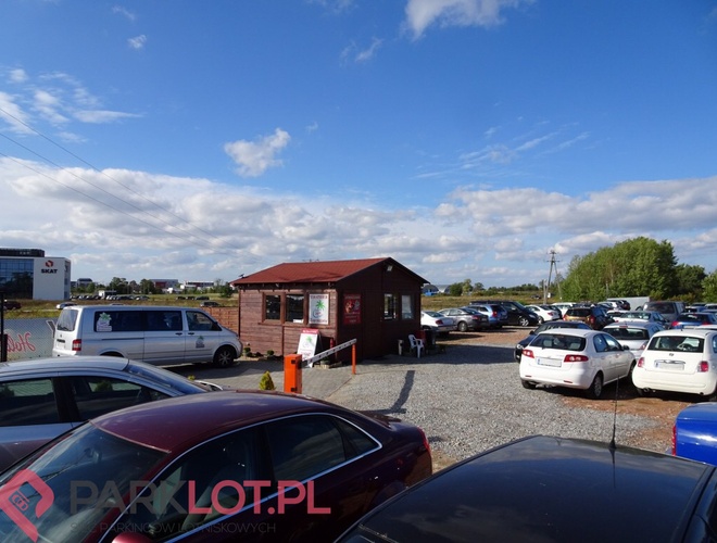 Holiday Parking Gdańsk - Na żwirowym placu stoją samochody. Obok znajduje się drewniany budynek. W tle widać ogrodzenie.
