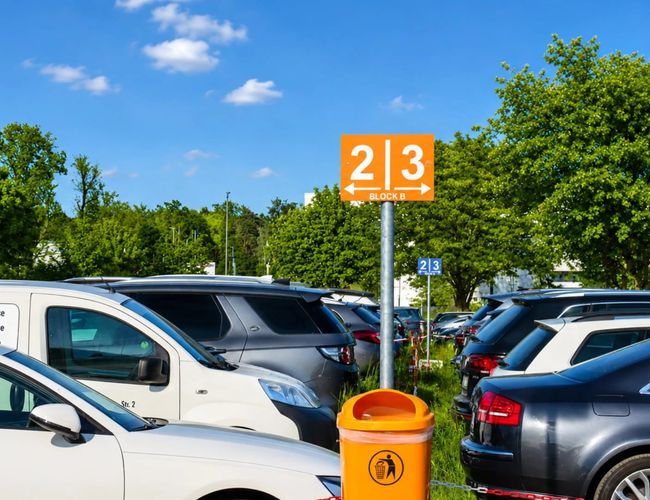 Parking myParkplace - Na żwirowym podłożu stoi duży bus. Obok znajduje się kolorowy plakat promocyjny z palmami i napisem. Dookoła są drzewa i samochody.
