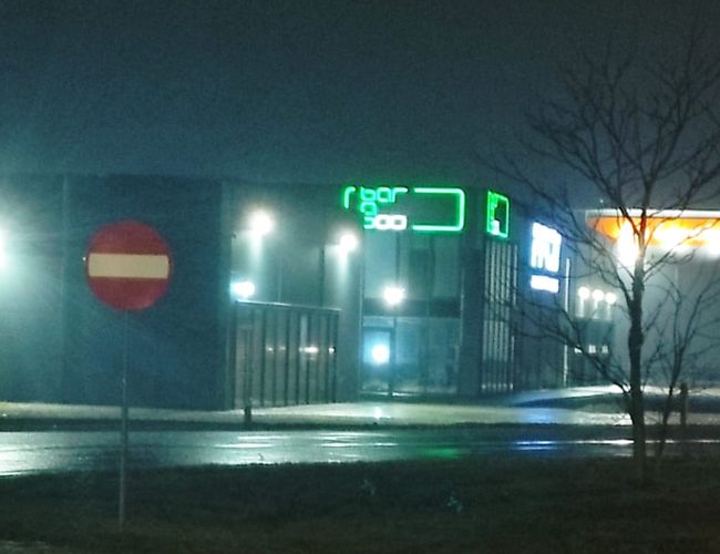Q-Parking — W tle neonowy napis nad budynkiem. Na pierwszym planie droga z zakazem wjazdu. Wieczorna sceneria z oświetleniem ulicznym.