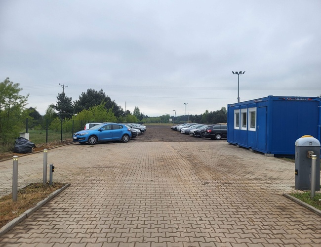 Primer Estacionamiento - En la plaza hay coches aparcados. Al lado hay un contenedor azul. El terreno está pavimentado con adoquines.