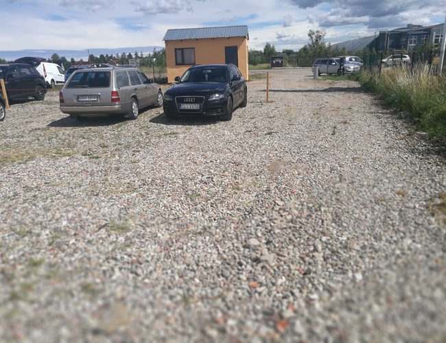 Parking P8 - Na żwirowym placu stoją samochody. W tle widać budkę. Parking jest na otwartej przestrzeni.