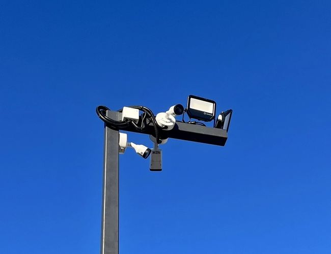 Parking Blisko Lotniska - Na zdjęciu latarnia z kamerami monitoringu. Niebo jest bezchmurne i niebieskie. Latarnia jest metalowa i wysoka.