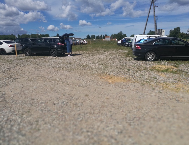 Parking P8 - teren utwardzony, kilka zaparkowanych samochodów. Jedna osoba przy ciemnym aucie. Pogoda jest słoneczna.