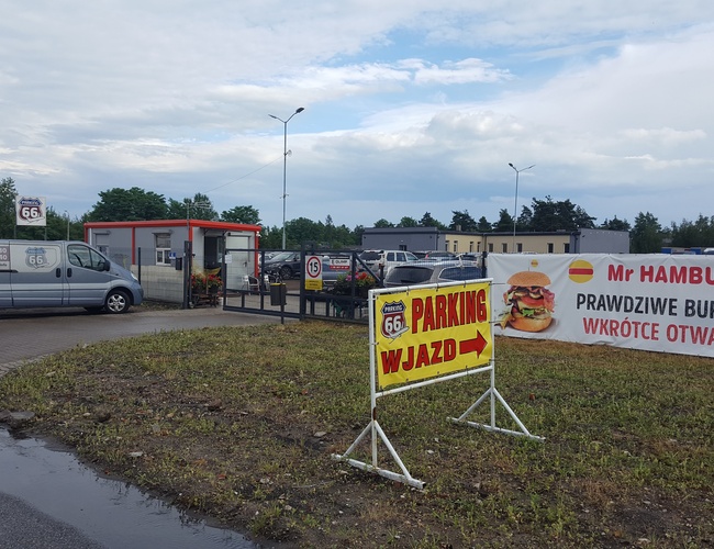 AAP-66 Parking – teren otwarty z bramą wjazdową. Zwisają reklamy burgerów. Dwa samochody stoją po lewej stronie.