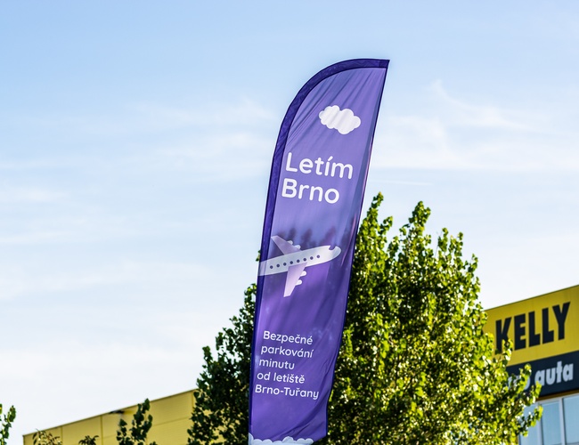 Parking Letim Brno – na zdjęciu widoczna jest fioletowa flaga reklamowa. Znajdują się na niej napisy informacyjne oraz grafika samolotu. W tle widać drzewo i fragment budynku.