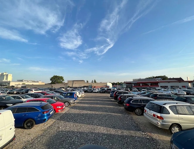 Parqueo de Aeropuerto Parktiger 2 - Gran aparcamiento no pavimentado con numerosos coches. El área está abierta y iluminada. Se pueden ver edificios al fondo.