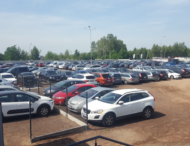 AAP-66 – Duży parking pełen samochodów. Teren jest ogrodzony. Otoczenie to drzewa i zieleń.