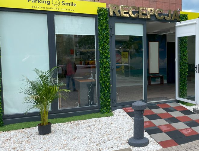 Parking Smile – Widać recepcję z dużym napisem. Na zewnątrz jest roślina i ozdobne płyty.