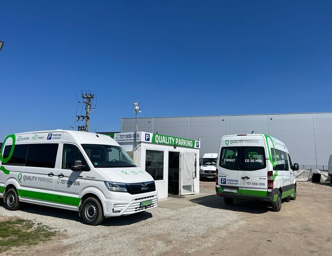 Q-Parking - Dwa białe busy stoją obok siebie. Obok znajduje się budynek z napisem "Quality Parking". W tle widać ogrodzenie.