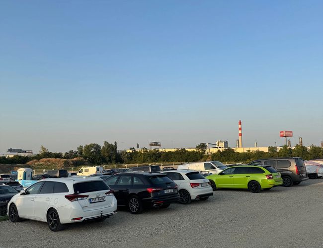 Rząd samochodów zaparkowanych na parkingu. Widoczna utwardzona nawierzchnia i drzewa w tle.