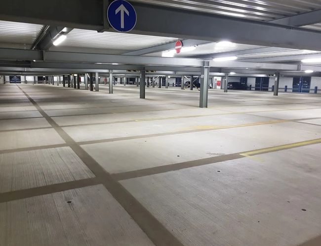 Przestronne wnętrze parkingu wielopoziomowego z szerokimi pasami ruchu, wyznaczonymi miejscami postojowymi oraz czytelnym oznakowaniem kierunkowym.