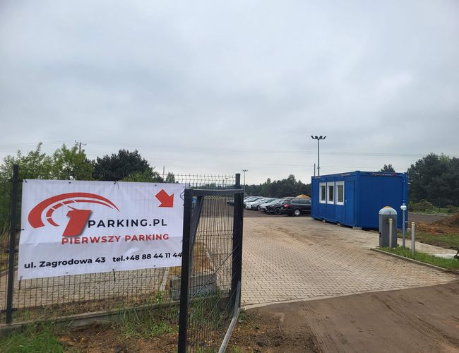 Primer estacionamiento - El terreno está cercado y pavimentado. A la derecha hay un contenedor azul. Al fondo se ven coches estacionados.