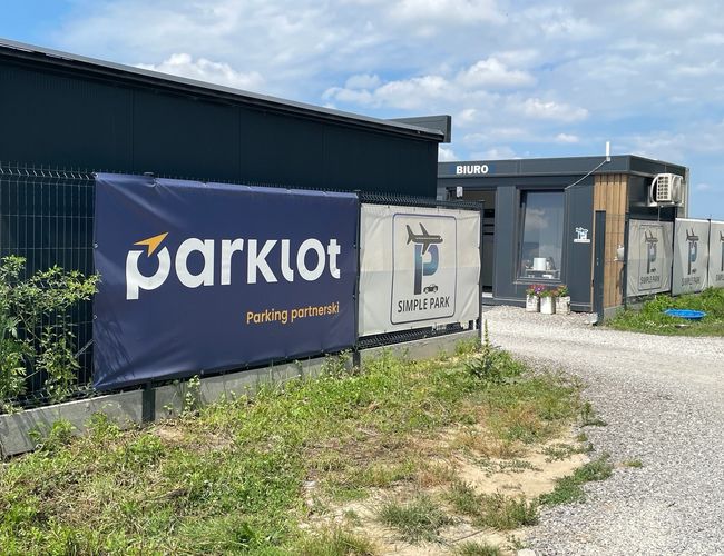 Parking Simple Park - Teren jest ogrodzony i oświetlony. Zaparkowany jest ciemny samochód. Obok znajduje się tablica informacyjna.