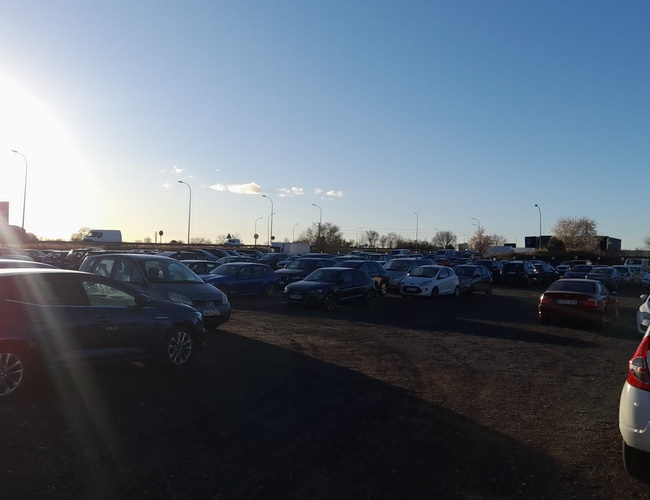 Parkovisko Pleyades Valet - Parkovisko plné áut v slnečný deň. Na zemi leží jemný štrk. Pouličné lampy stoja po stranách.