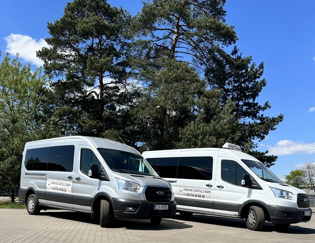 Estacionamiento Inicia con Nosotros - Dos furgonetas blancas están en una superficie pavimentada. Al fondo hay árboles altos.