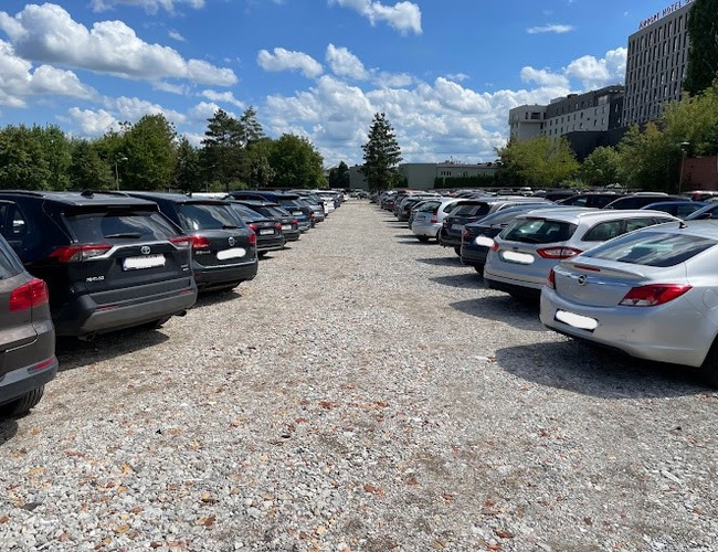 Parking Smile - Żwirowa powierzchnia z zaparkowanymi samochodami po obu stronach. Otoczony drzewami i budynkami. Bez ogrodzenia.