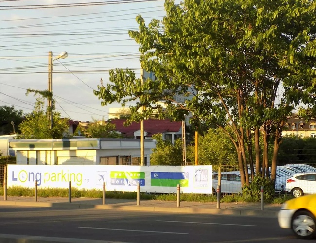 Wejście na parking „Long Parking”, ogrodzenie z reklamami i drzewa w tle w słonecznym otoczeniu.