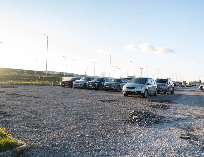 Easy Parking Express - Parking jest żwirowy i otwarty. Samochody są zaparkowane w równych rzędach. Lampy zapewniają oświetlenie.