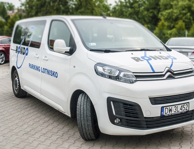 Parking Rondo - Biały van z logiem parkingu stoi na kostce. W tle są inne zaparkowane samochody i zielone drzewa.
