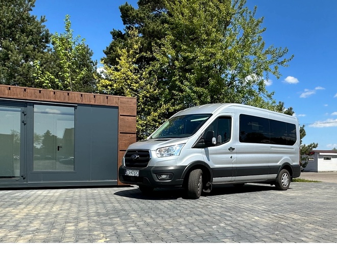 Empieza con nosotros – La minibus está al lado de la oficina contenedor. Alrededor crecen árboles, y el cielo es soleado.