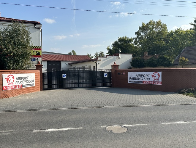 Estacionamiento del aeropuerto 500 - Puerta de entrada rodeada de un muro de ladrillos. Señales y tablones informativos a los lados. Al fondo, árboles y edificios.