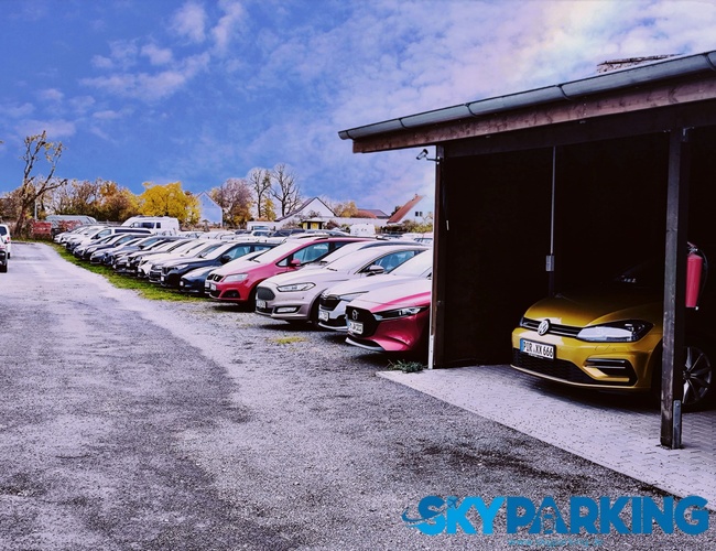 Długi rząd aut zaparkowanych na otwartym parkingu o utwardzonej nawierzchni, obok którego stoi niewielka wiata; w tle widoczne są drzewa oraz jasne niebo.