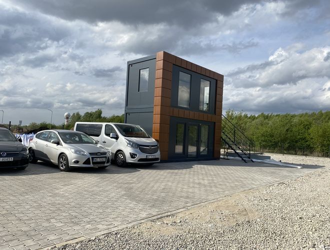 SkyWay Parking – Na parkingu stoją samochody osobowe i van. Obok znajduje się piętrowy budynek zewnętrzny. Teren jest utwardzony i ogrodzony.