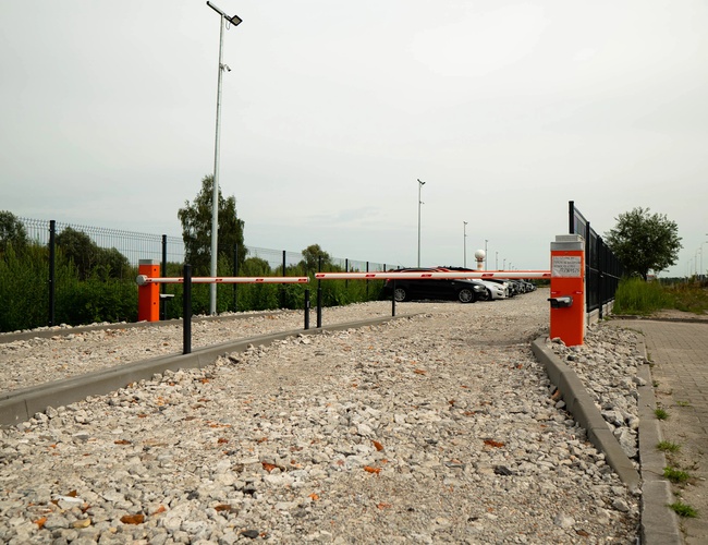 Easy Parking Express – Vjazd so závorou na štrkovom povrchu. Parkovisko je ohradené a osvetlené lampami. V pozadí stoja zaparkované autá.