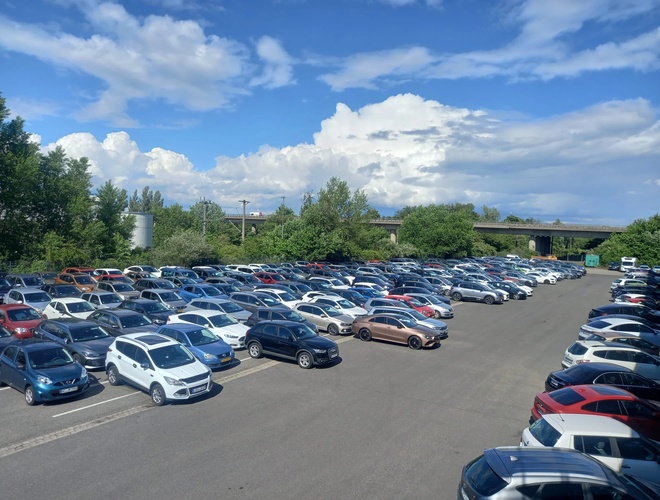 Duży parking pełen samochodów osobowych pod błękitnym niebem z nielicznymi chmurami, w tle drzewa i budynki.