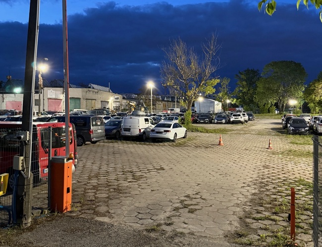 Widok wjazdu ze szlabanem na parking nocą, dobrze oświetlony plac.