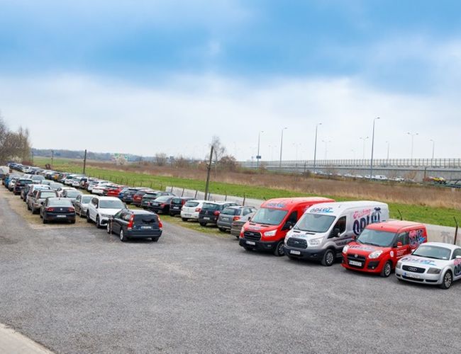 Parking Odlot - Na żwirowej powierzchni stoi wiele samochodów. Parking jest ogrodzony. W oddali widać drogę i latarnie.