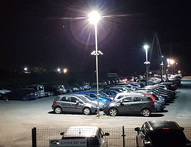 Parking MTM – Dobrze oświetlony plac wypełniony samochodami. Widoczne latarnie i ogrodzenie. Nocna sceneria z kilkoma światłami w tle.