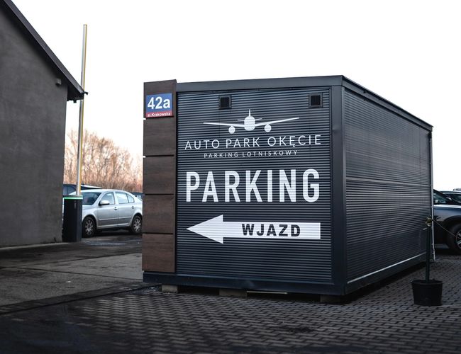 Auto Park Okęcie - kontener z napisem „Parking, Wjazd” znajduje się na brukowanej nawierzchni. W tle widoczne są samochody i budynki.