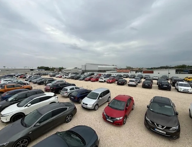 Duży parking wypełniony zaparkowanymi samochodami osobowymi na żwirowym placu, widok z lekkiej wysokości.