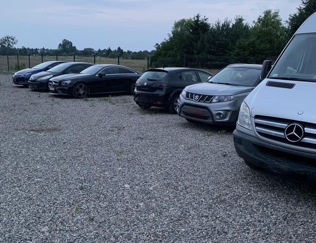 A1 Parking Max – Na żwirowym placu stoi kilka samochodów. Teren jest ogrodzony siatką. W tle widoczne są drzewa i otwarta przestrzeń.
