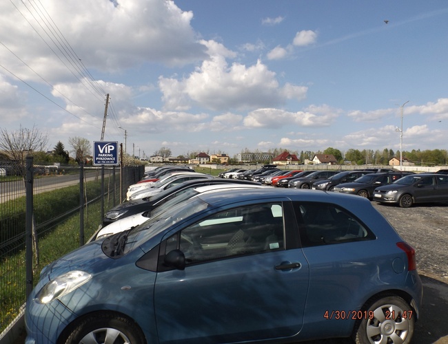 VIP Parking – Na zdjęciu widoczny jest ogrodzony plac z samochodami. Obok parkingu biegnie droga. Na pierwszym planie jest niebieski samochód.