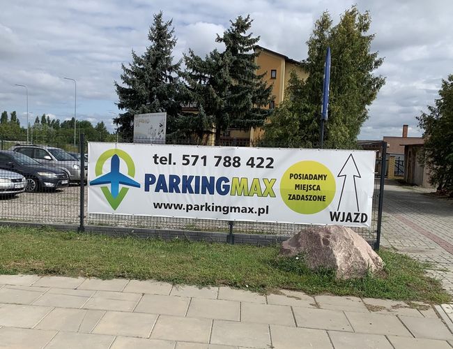 A1 Parking Max – Parking jest ogrodzony siatką. Na terenie stoją zaparkowane samochody. Widoczny jest duży billboard reklamowy.
