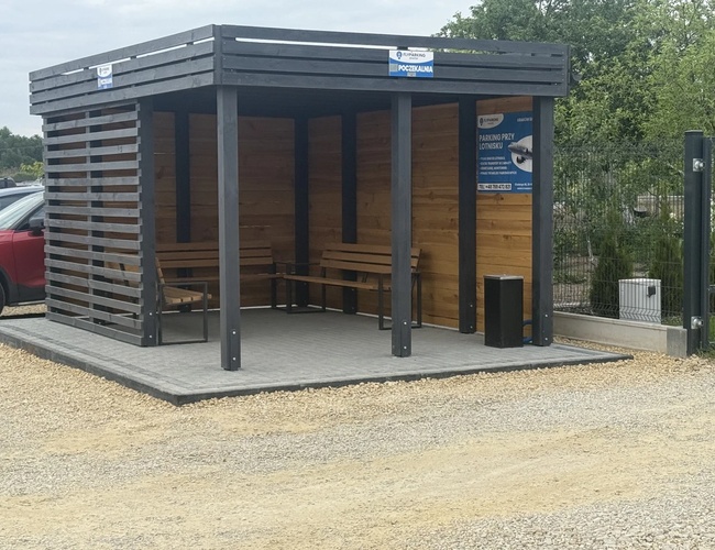 FlyParking - Drevená čakáreň s dvoma lavicami stojí vedľa parkoviska. Strecha chráni pred dažďom. Vedľa je zaparkované červené auto.