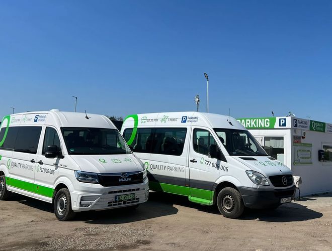 Q-Parking – Dwa białe busy stoją przed budką z napisem. Teren jest nieutwardzony, widoczne jest ogrodzenie z siatki.