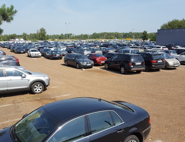 AAP-66 – Parking wypełniony samochodami osobowymi. Jasny, słoneczny dzień. Widoczna niska zabudowa w tle.