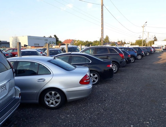 Parkovisko P7 - Automobily zaparkované na štrkovom teréne pri budovách. Pozemok je nezaistený s viditeľným osvetlením ulicných lámp.