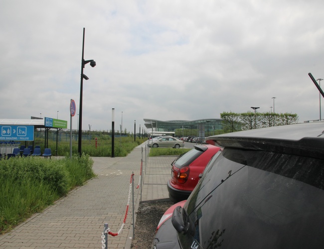 Oficjalny Parking Lotnisko Wrocław Strachowice – zaparkowane auta obok terminala. Widać chodnik i oznaczenia wózków bagażowych.