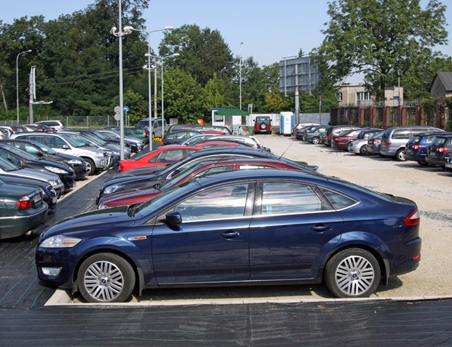 Parking Rondo - Na parkingu stoi wiele samochodów. Teren jest otwarty i oświetlony. W tle rosną drzewa.