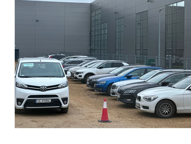 Holiday Car Park - Na żwirowym parkingu stoi rząd samochodów osobowych. Otoczony jest nowoczesnym budynkiem z dużymi oknami.