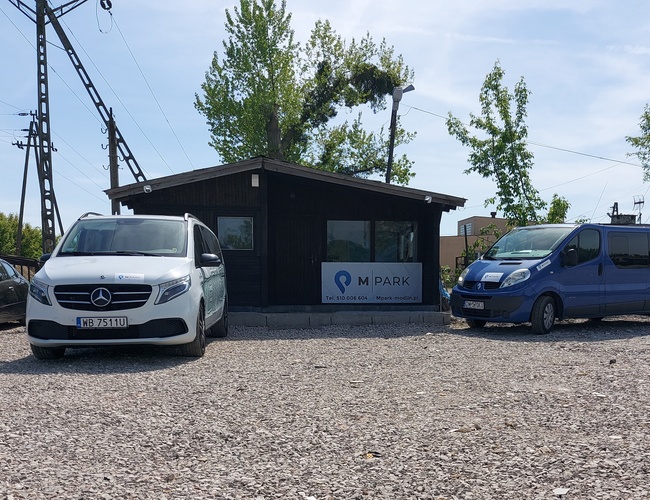 Parkovisko MPark - Pred budovou sú zaparkované tri autá. Pozemok je pokrytý štrkom a obklopený stromami.